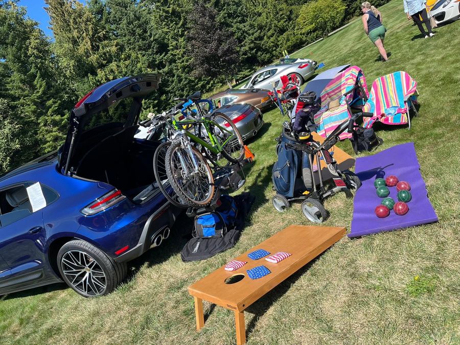 Porsche trunk open with bikes, golf clubs, cornhole, and bocce ball set up on the lawn